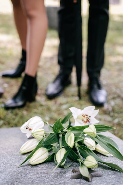 A bouquet of white lilies with partially opened petals and green leaves, arranged with elongated stems and buds, resting on a stone surface. In the background, a blurred scene of a person in formal bl