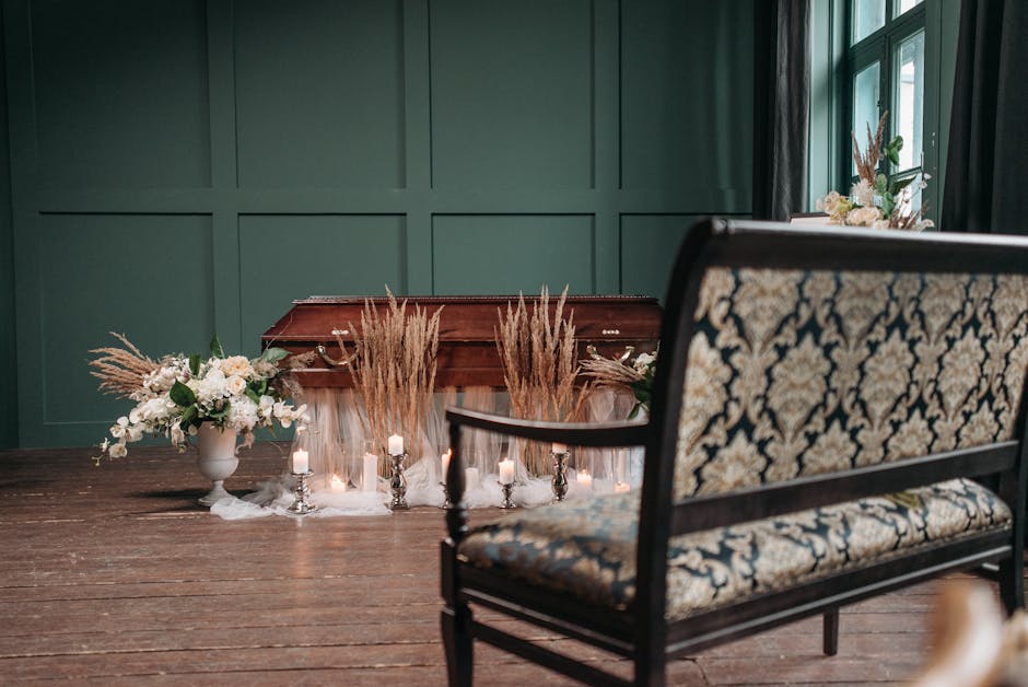 A wooden casket is positioned against a dark green paneled wall in a softly lit room, with natural light filtering through a nearby window. In front of the casket, there are floral arrangements includ