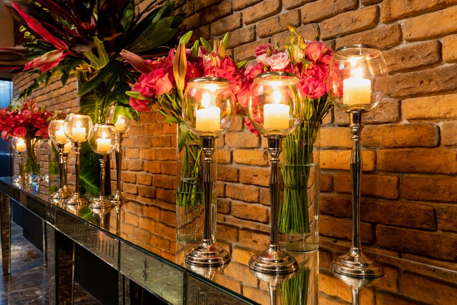 A series of elegant floral arrangements featuring large glass vases filled with vibrant pink lilies and roses, complemented by dark green foliage, set along a reflective black surface against a warm, 