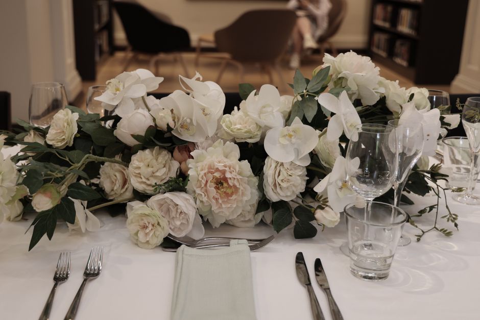 A long, luxurious floral arrangement featuring predominantly white flowers, including large peonies, orchids, and roses, accented with lush green foliage, arranged in a cascading style across a white 