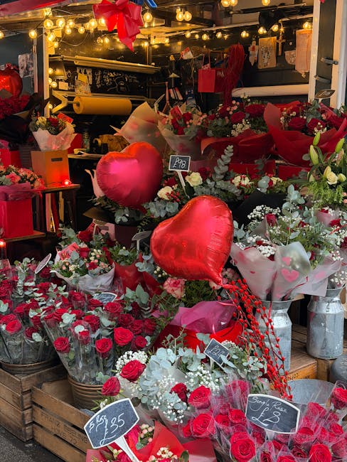 A vibrant display of floral arrangements at a flower shop, featuring predominantly red roses, white blooms, and greenery, arranged in buckets and baskets. The display includes heart-shaped balloons an
