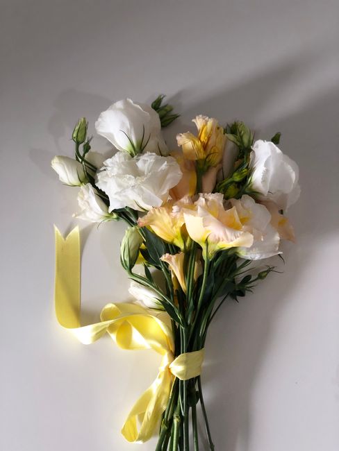 A bouquet arrangement of fresh white and pale yellow lisianthus flowers, with ruffled petals and slender green stems, tied with a yellow satin ribbon. The bouquet is laid on a neutral background, show