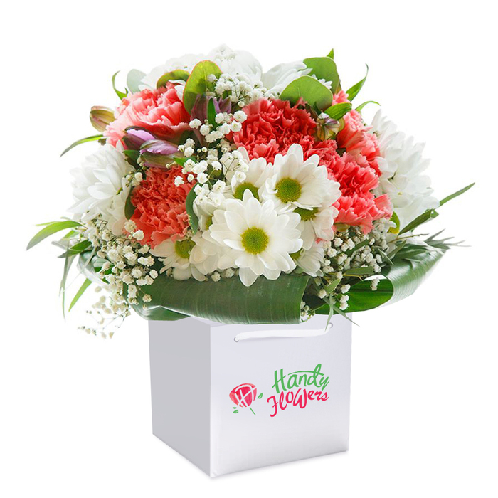 A beautifully full and vibrant bouquet, arranged with flair by experienced Totteridge florists, is poised in a pristine, square white gift bag-its presentation as crisp and inviting as the air around Totteridge Common on a bright morning. The lush cluster of blooms is dominated by coral, peach, and classic red carnations, each distinguished by their layered, ruffled petals that catch the light and evoke both softness and abundance. Embraced within the bouquet's rounded silhouette are crisp white daisies, their lime-hued centers popping like bursts of spring against the cheerful coral shades. Wisps of baby's breath are artfully interwoven throughout, offering a delicate, ethereal texture that lifts the arrangement and imparts a dreamy atmosphere. Subtle buds showing hints of purple nest along the edges, providing a sense of depth and gently hinting at blossoms yet to open-a nod to the ever-changing faces of Totteridge's gardens and wild spaces. Encompassing the entire collection are broad, glossy, deep green leaves, forming a generous collar that frames the bouquet and adds a sense of structural grace. The sturdy, modern gift bag, fitted with satin ribbon handles for easy carrying, suggests a ready-to-gift intention-perfect for deliveries to local addresses from High Road to Dollis Brook. Shot against a flawless white backdrop, every detail, from petal to leaf, is illuminated, celebrating freshness, expert arranging, and the refined simplicity Totteridge residents adore in their floral gifts for birthdays, anniversaries, or any day that calls for a little extra joy.